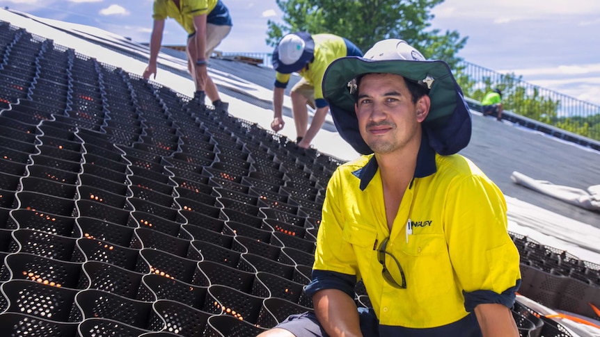 A man in high-visibility gear and sun shaded construction hat sits with plastic grid structures on a sloping roof