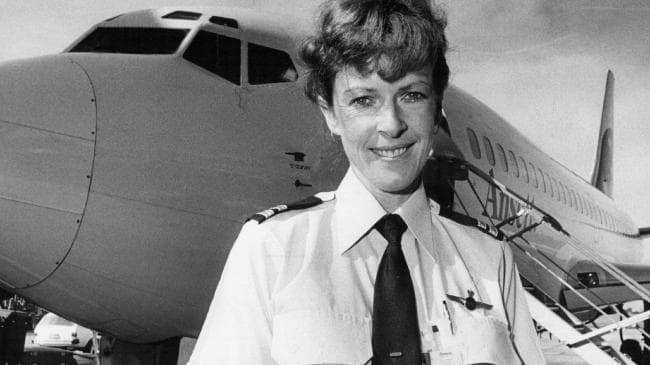 Black and white photo of smiling woman in pilot uniform standing in front of grounded Ansett aeroplane.