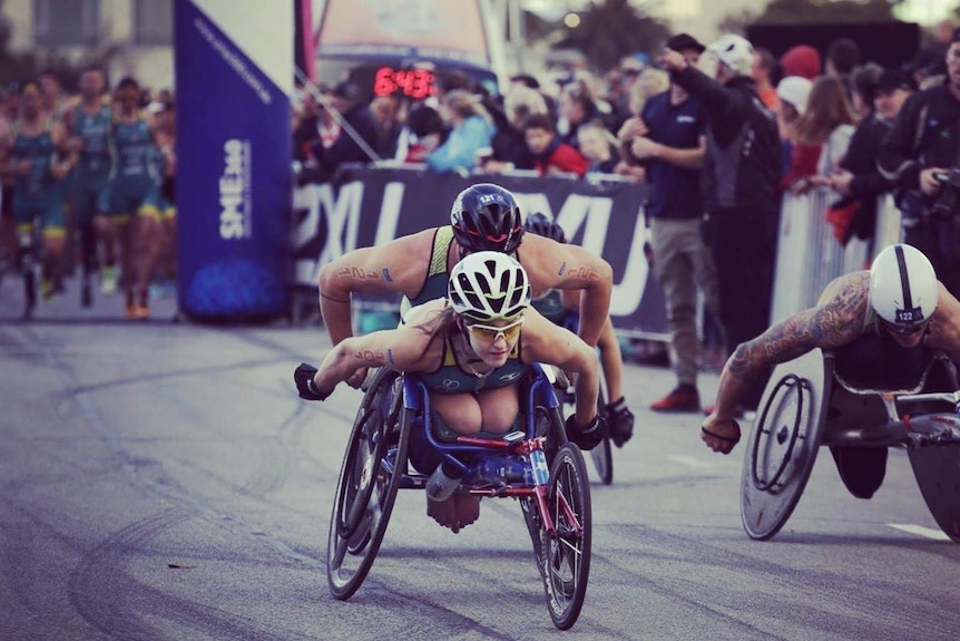 A woman races in a wheelchair with two other athletes in front of a crowd.