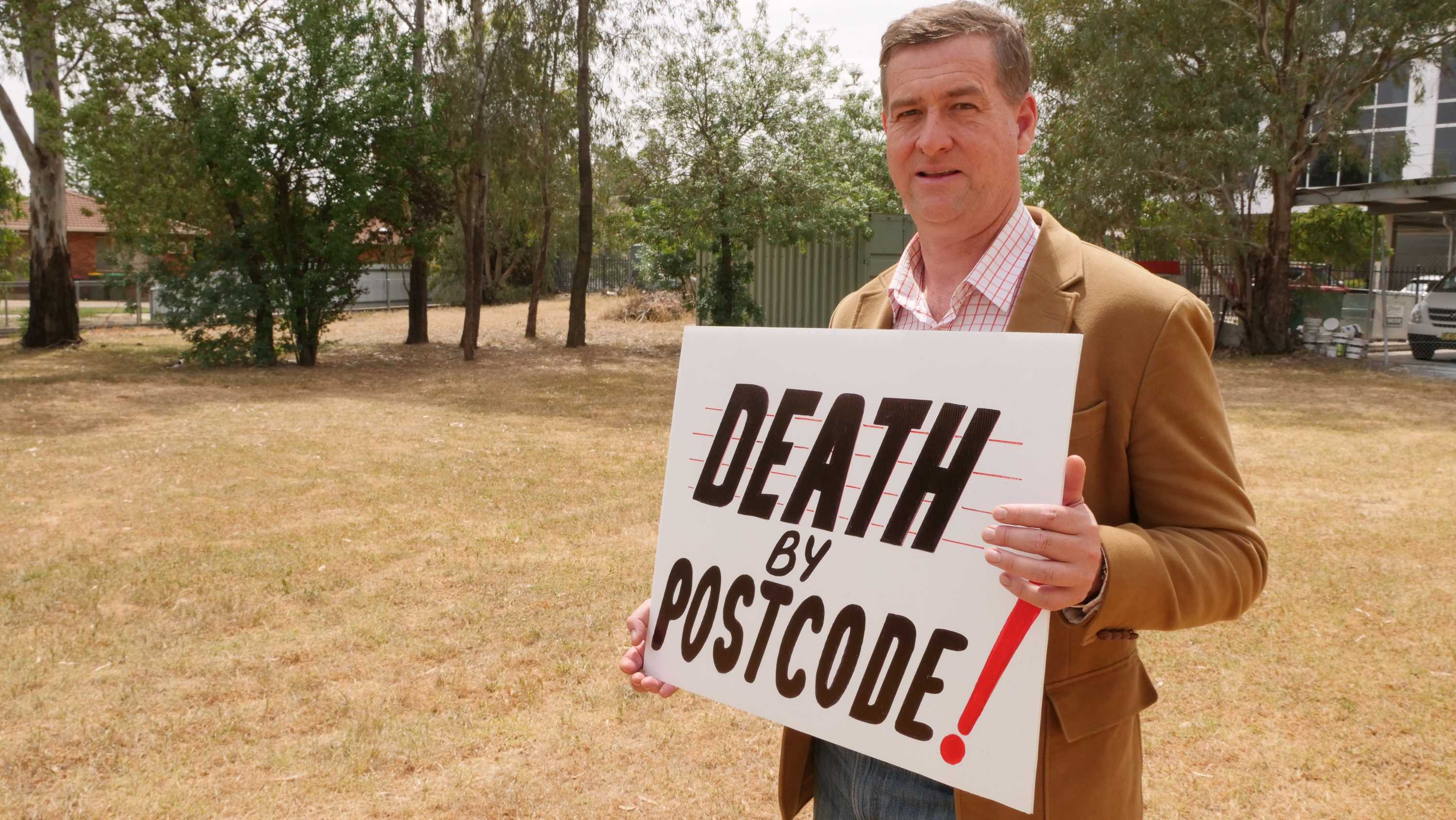 Man standing in empty block holding a sign saying death by postcode