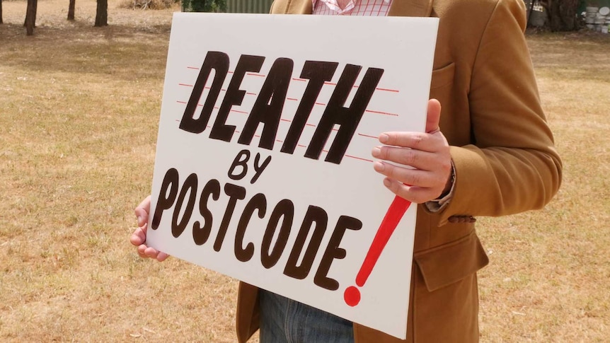 Man standing in empty block holding a sign saying death by postcode.