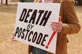 Man standing in empty block holding a sign saying death by postcode