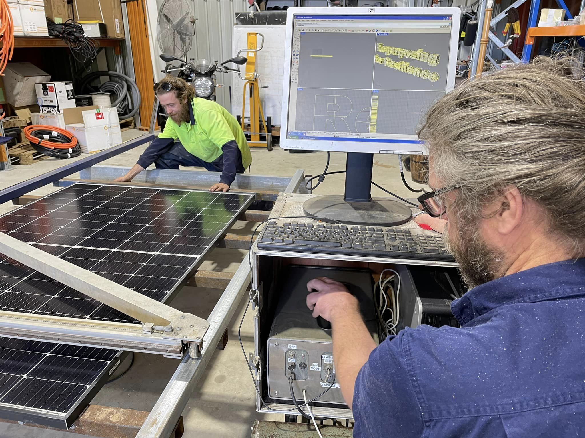 Two men work on a computer and a cutter with a large solar panel inside a shed.