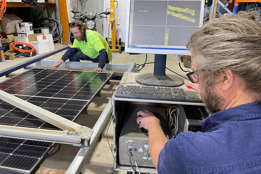 Two men work on a computer and a cutter with a large solar panel inside a shed.