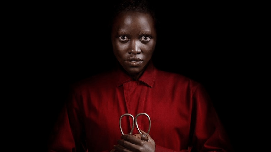 A black woman in a red jumpsuit stares at the camera, holding a pair of gold scissors.