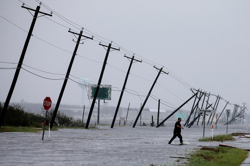 A man walks through flood waters and onto the main road as telephone poles fall down behind.