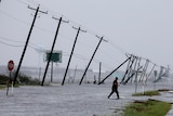 A man walks through flood waters and onto the main road as telephone poles fall down behind.