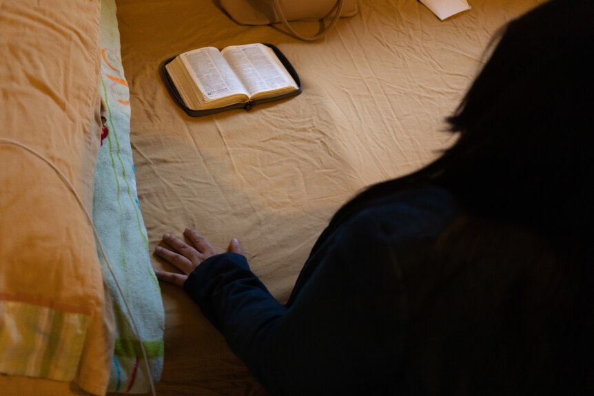 A woman puts her hands on a bed in front of an open Bible.