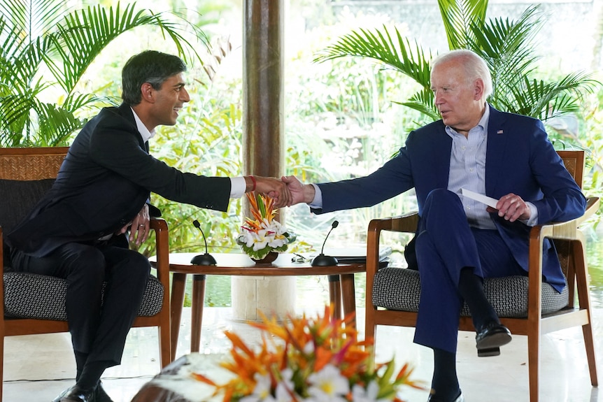 Sunak and Biden lean forward to shake hands from where they sit on wooden chairs side by side on a raised platform