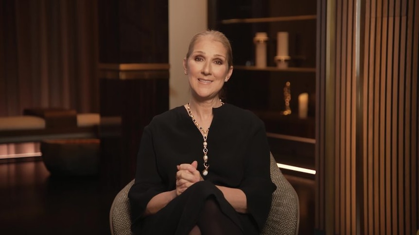 Celine Dion sitting on a chair in an office, wearing a black long-sleeved top and a gold necklace.