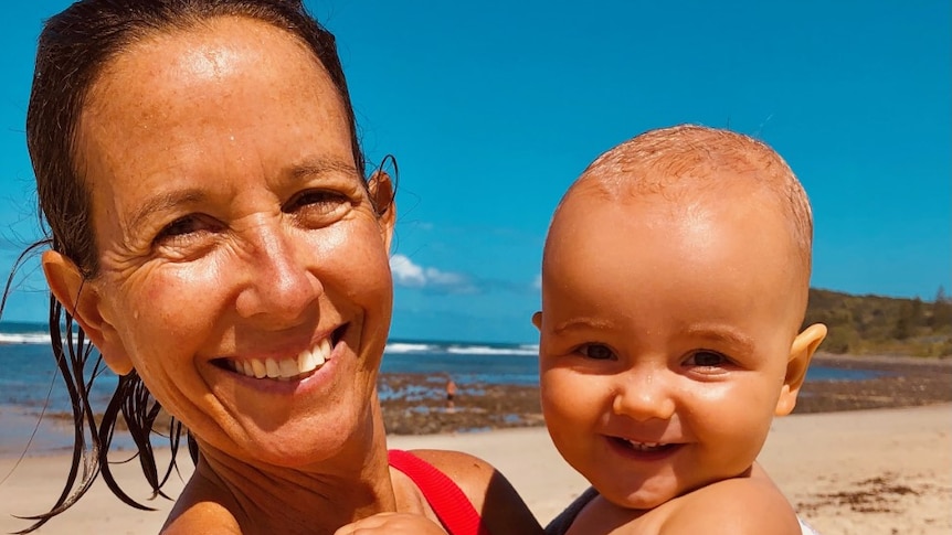 Woman grins on beach holding infant son