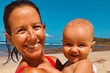 Woman grins on beach holding infant son