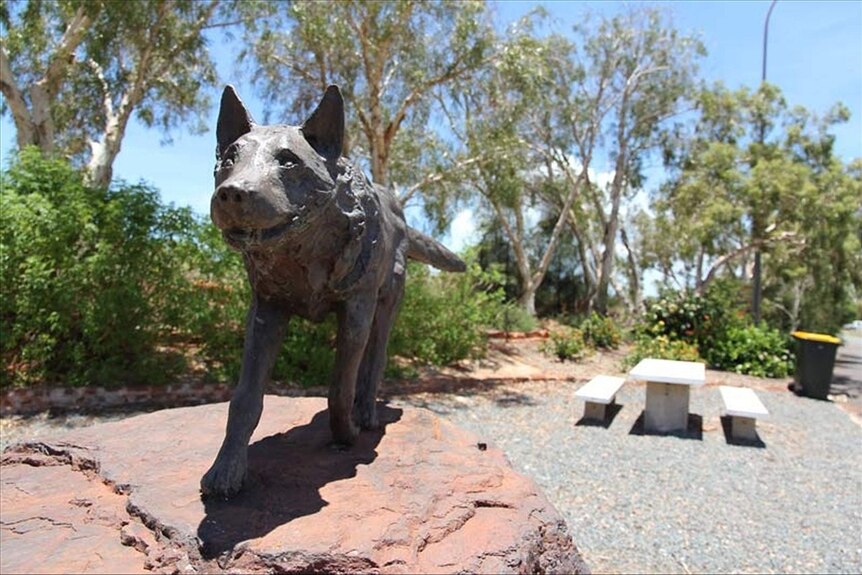 A statue of a dog on a red rock with greenery, a white table and seats and a bin in the background.