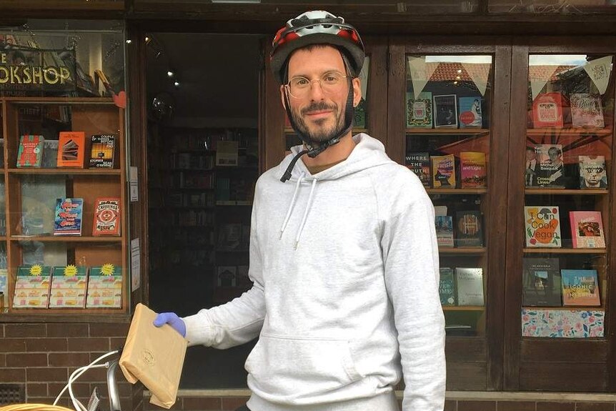 A man wears plastic surgical gloves and stands outside a bookshop with his bike holding a book in a paper bag.
