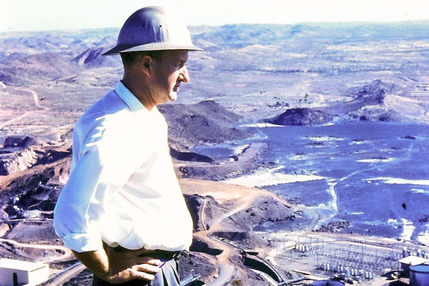 A man wearing a hard hat stands, hands on  hips, looking out into the distance. The photo is aged, from 1959.