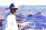 A man wearing a hard hat stands, hands on  hips, looking out into the distance. The photo is aged, from 1959.