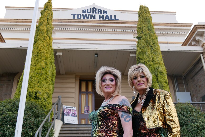 Two drag queens wearing gold sequin dresses and blonde wigs stand with two trees and a heritage building in the background. 