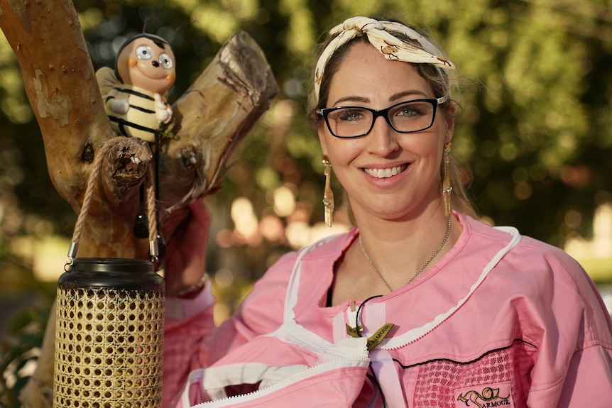 Alyson Shepherd in her pink beekeeping outfit without hat on, smiling next to a little figure of a bee