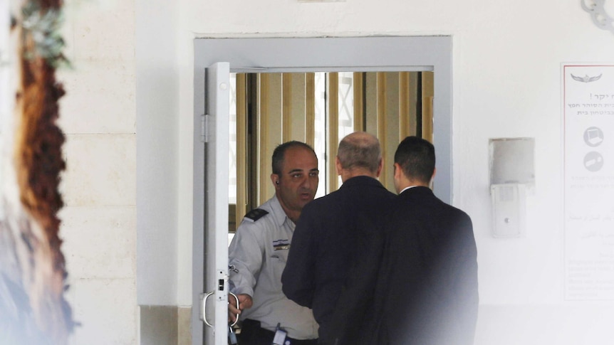 Ehmud Olmert is seen from behind as he is escorted by two people into a prison cell.