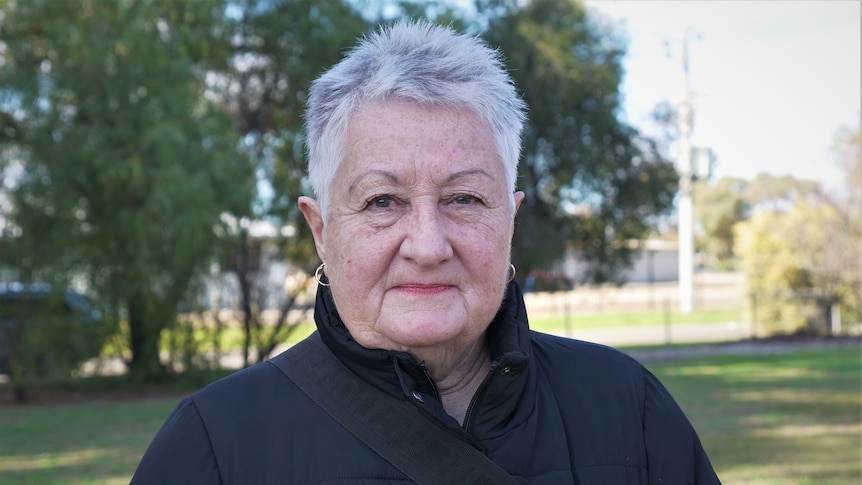 A woman with short grey hair looks into the camer with a neutral expression. She wears a black coat and gold earrings. 