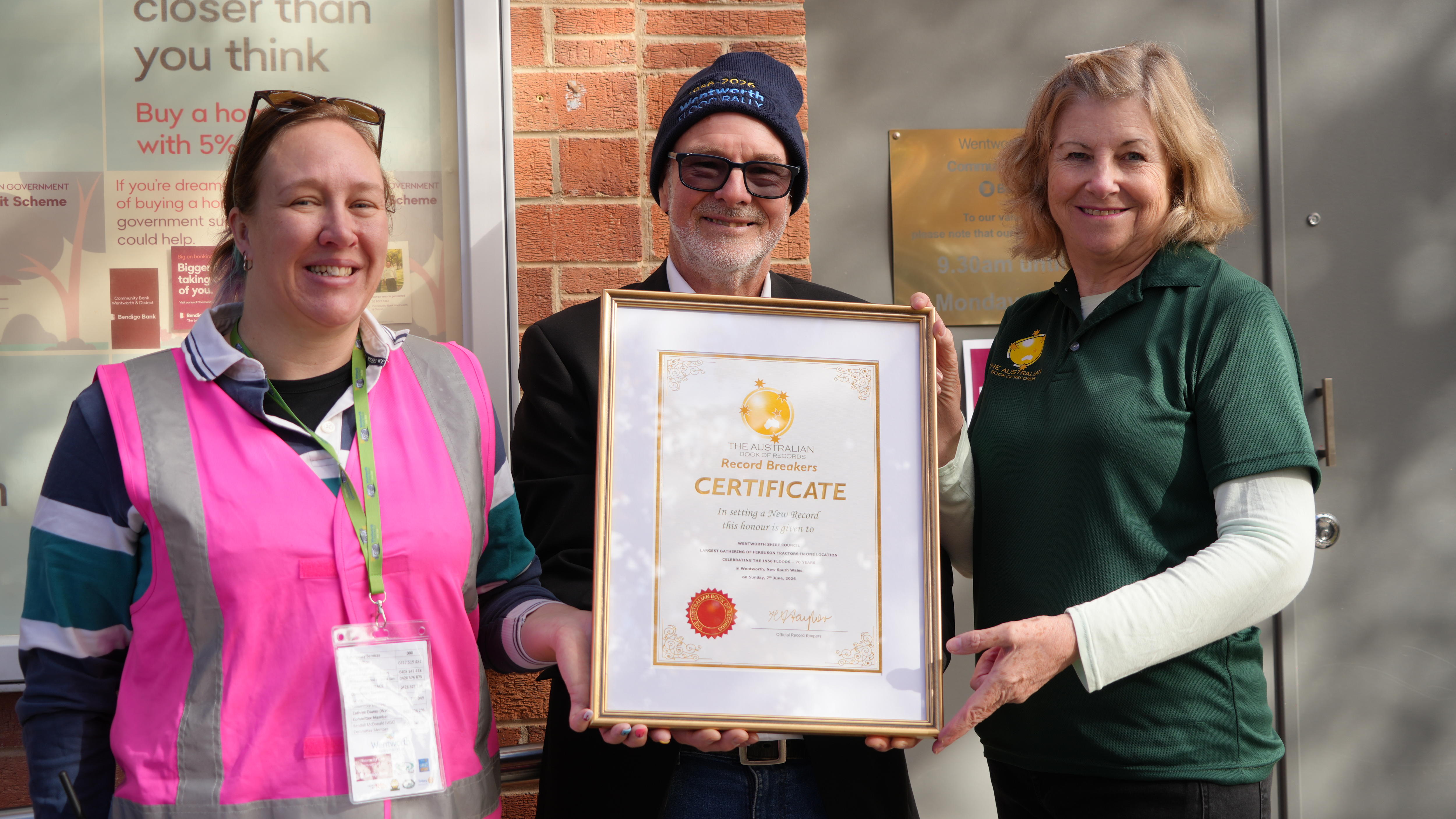 Un hombre con gafas de sol, perilla gris y gorra negra posa con un certificado junto a dos mujeres.