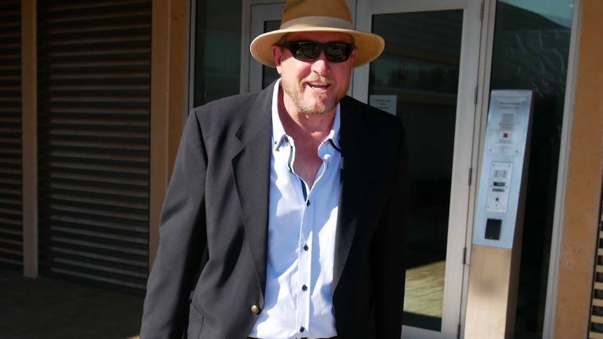A man in a shirt and jacket wearing a hat and sunglasses leaves a court building