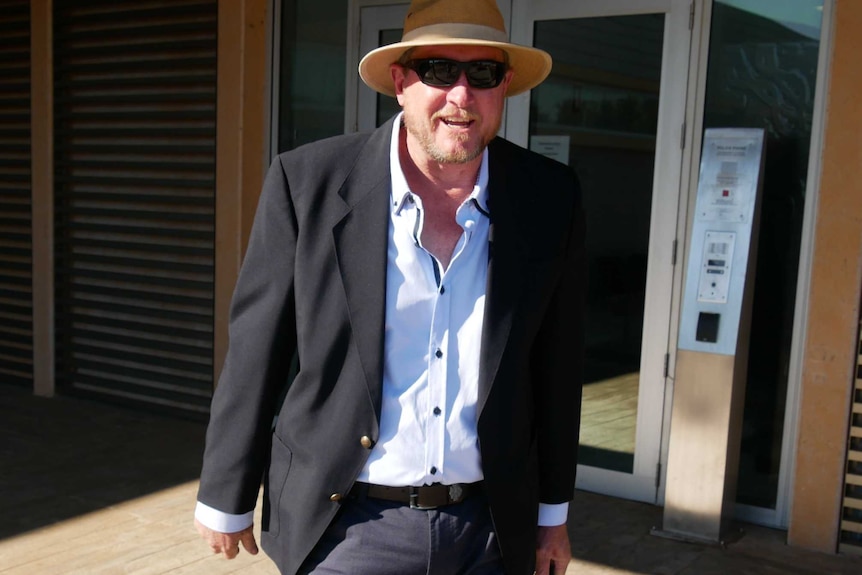 A man in a shirt and jacket wearing a hat and sunglasses leaves a court building