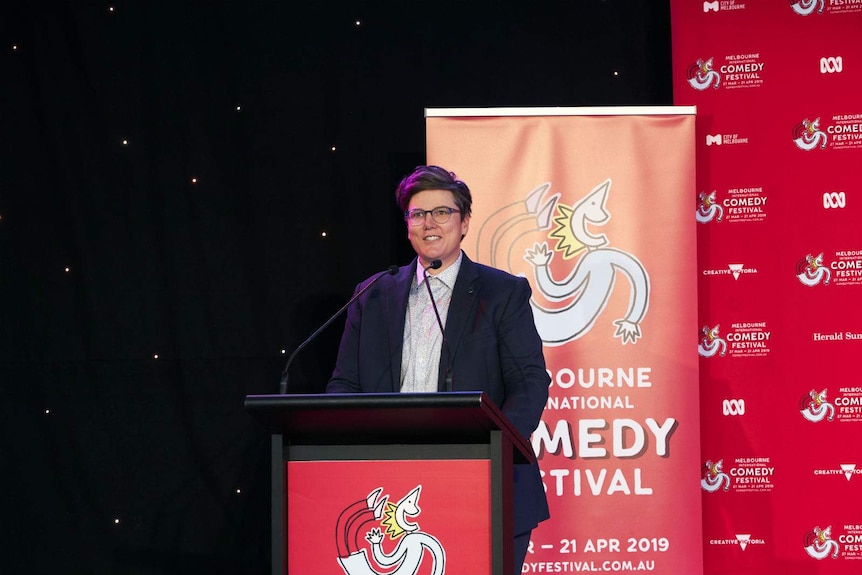 Gadsby stands smiling at a lectern, wearing a blue blazer, with Melbourne International Comedy Festival posters behind her.