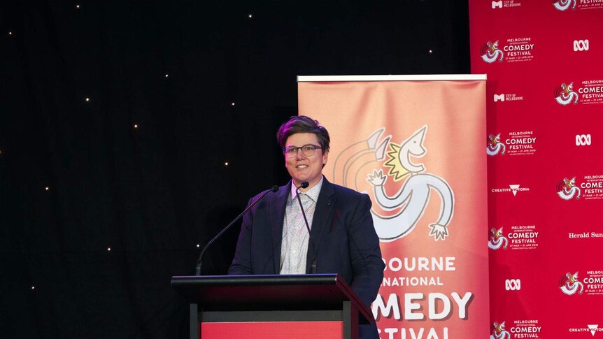 Gadsby stands smiling at a lectern, wearing a blue blazer, with Melbourne International Comedy Festival posters behind her.