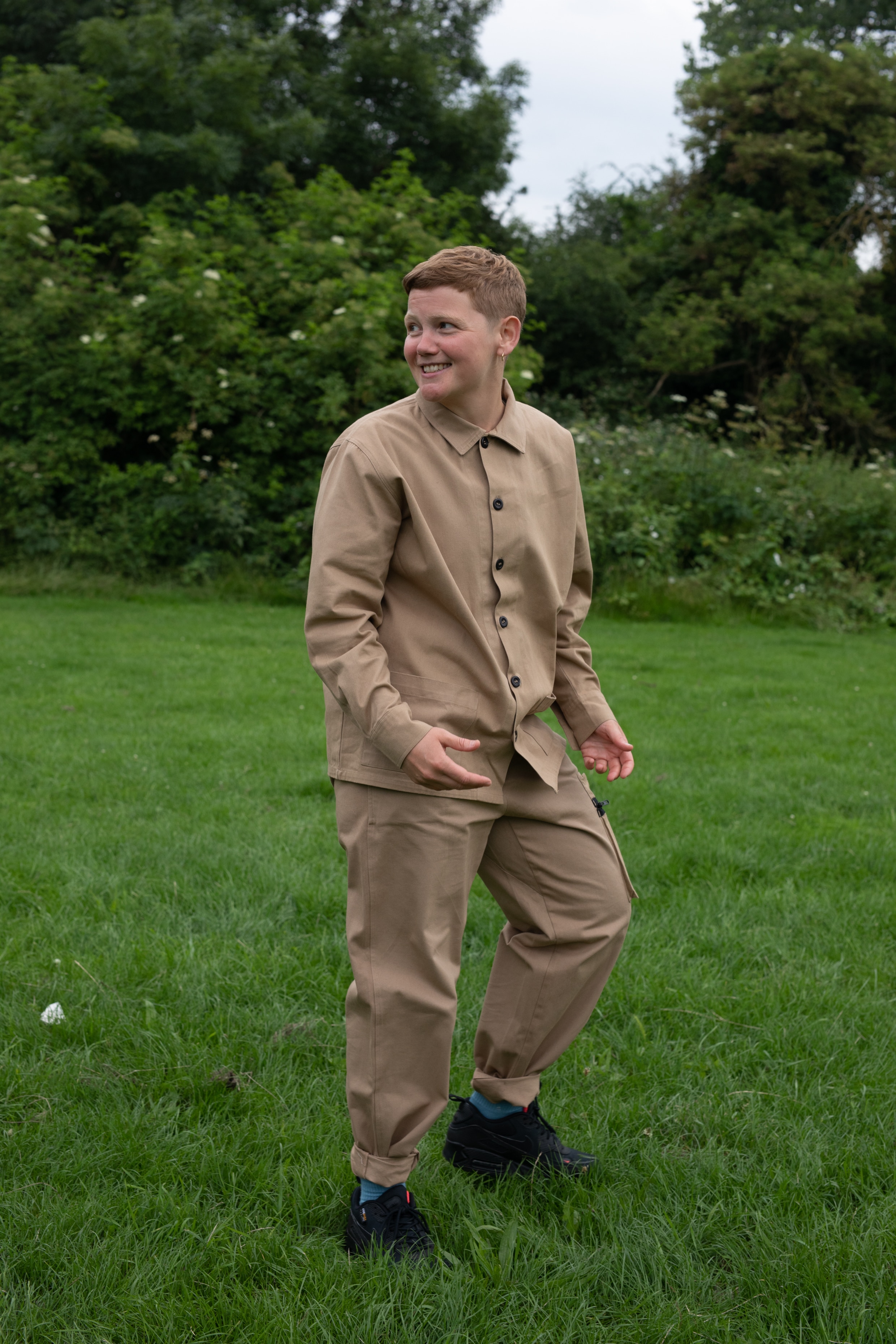 A white non-binary person with short sandy hair wears a long-sleeved khaki shirt and pants in a luscious grassy park.