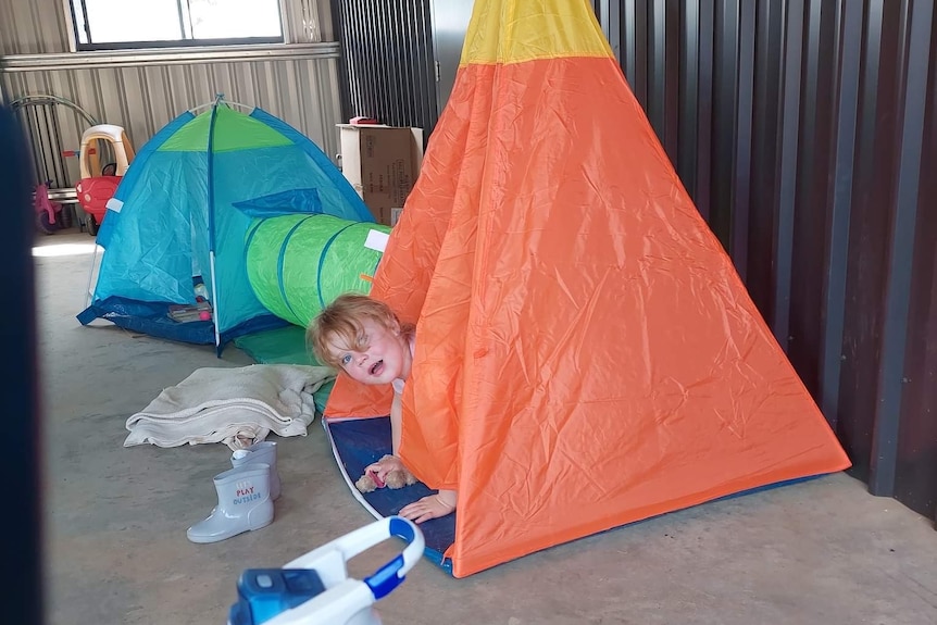 A toddler sits in a tent with her head poking out, placed around another tent and play items.