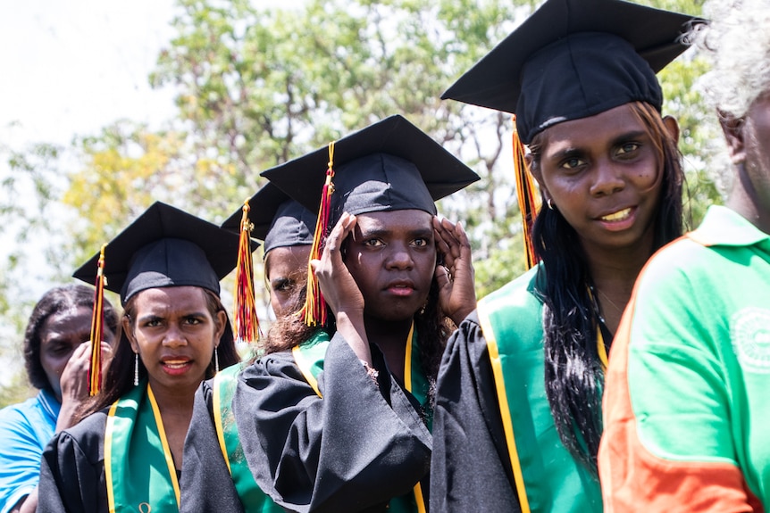 A row of young indigenous women dressed in graduation gowns stand outside under the sun.