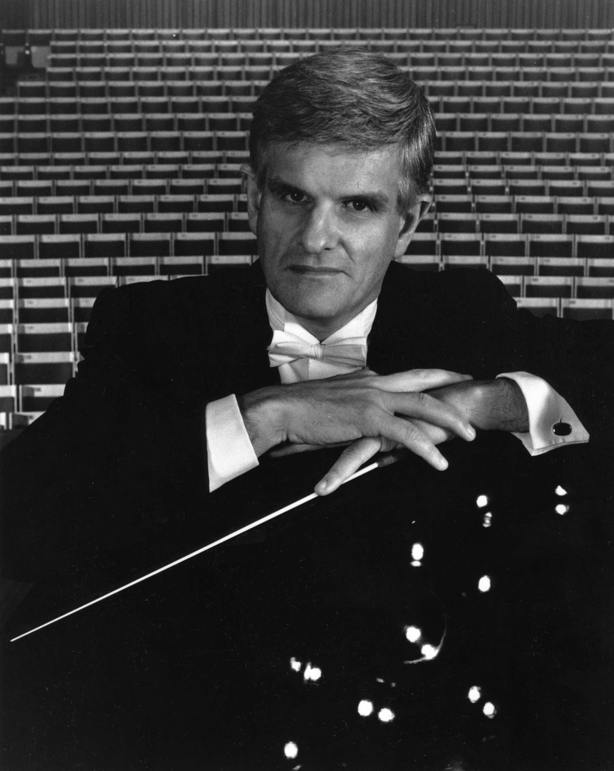 A black and white photo of a grey-haired white man wearing a formal black suit and holding a conductor's baton.
