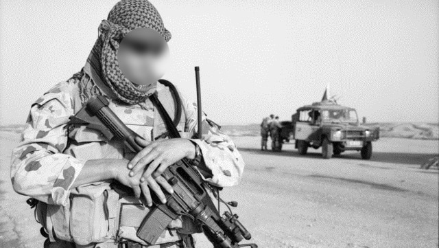 A black and white photo of a man in military clothing holding a gun. His face is blurred and there are people behind him.