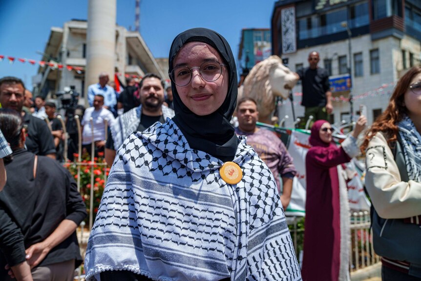 A young girl wearing a black and white kaffiyeh and glasses smiles among a crowd of people.