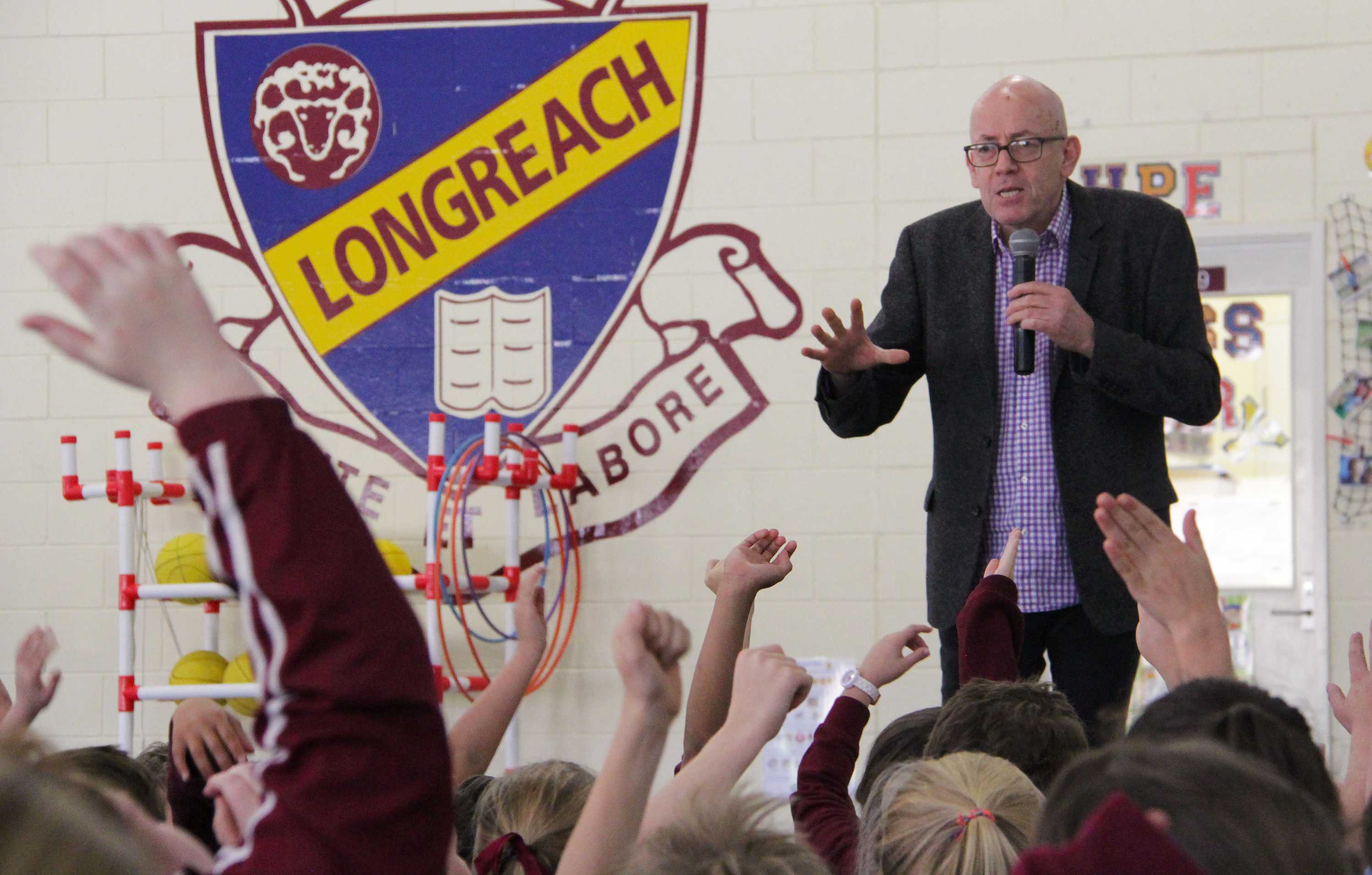 A man, gesturing with his hands, speaks to a large group of children with their hands raised to ask questions.