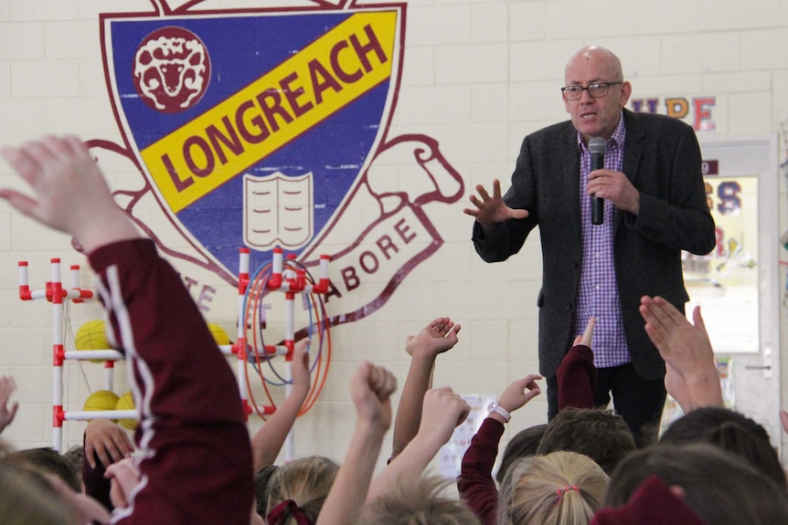 A man, gesturing with his hands, speaks to a large group of children with their hands raised to ask questions.