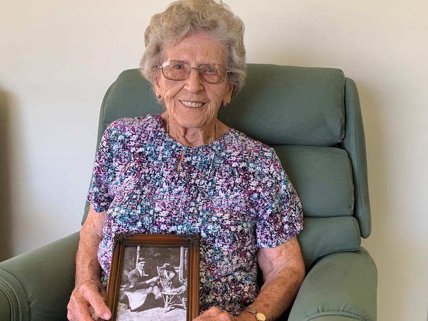 An elderly woman with grey hair wearing a floral shirt sitting in an armchair holding a framed photo of her father and Bluey.