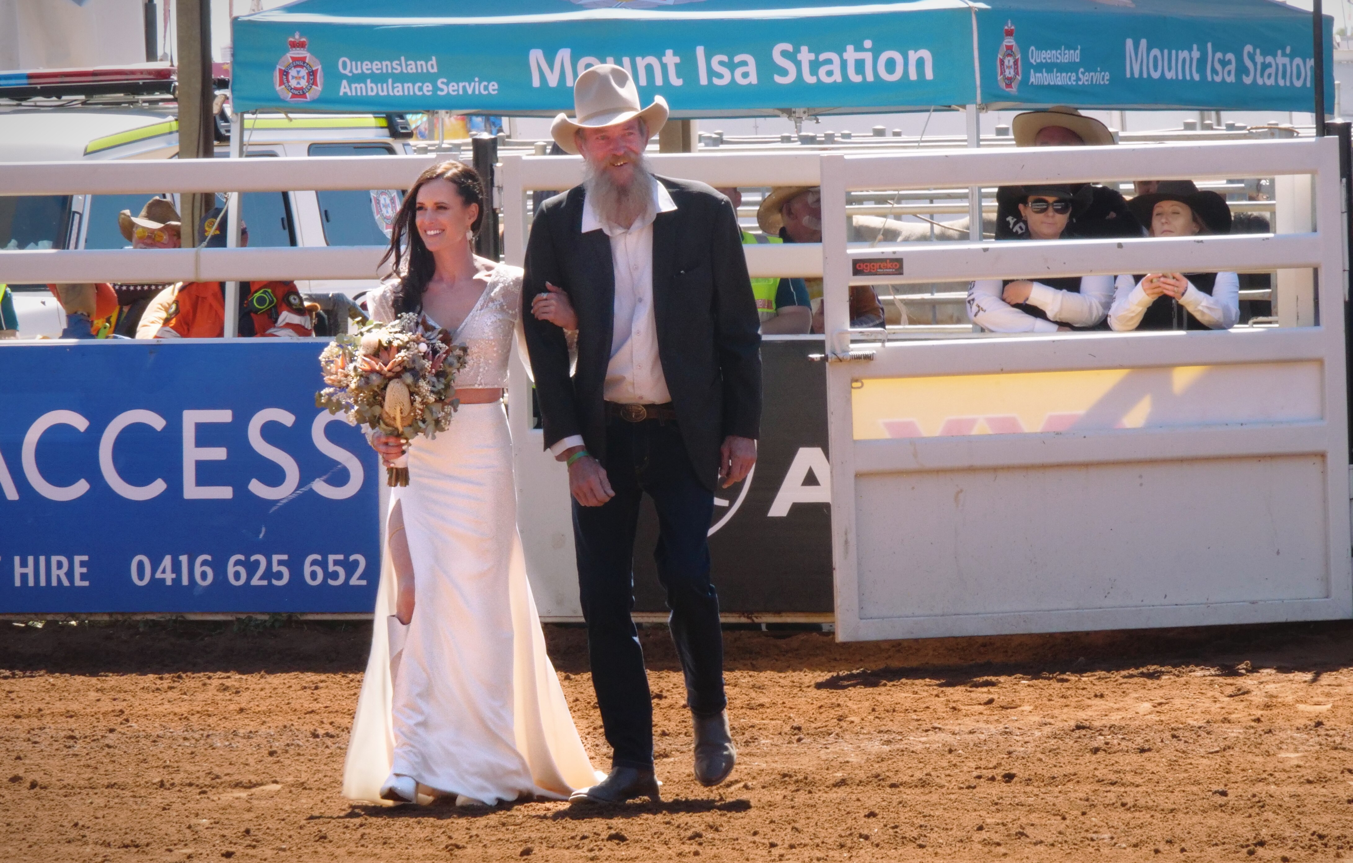 A bride holds a bouquet and walks through metal gates onto red dirt.  She's accompanied by a man in a suit and cowboy hat.