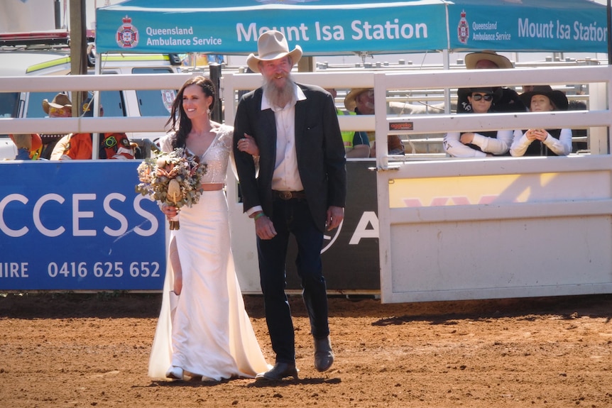 A bride holds a bouquet and walks through metal gates onto red dirt. She's accompanied by a man in a suit and cowboy hat.