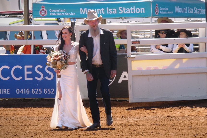 A bride holds a bouquet and walks through metal gates onto red dirt.  She's accompanied by a man in a suit and cowboy hat.