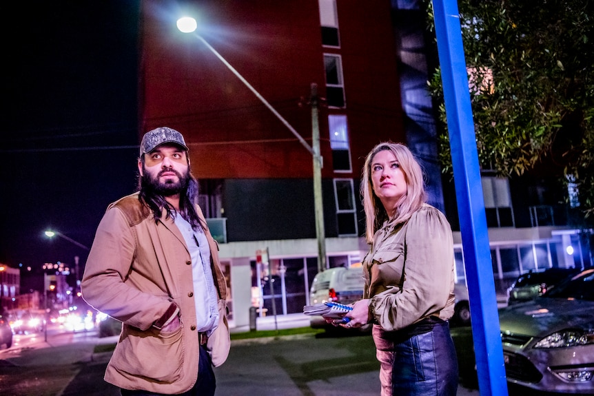 Under a bright streetlight, a man and a woman stand in front of a modern apartment block and look into the distance