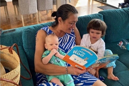 A woman sits on a teal couch, reading to a little boy and a baby.