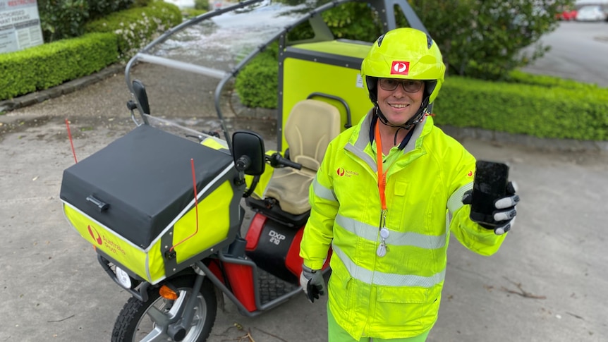 A man wearing hi-vis smiles at the camera while holding up a mobile phone. 