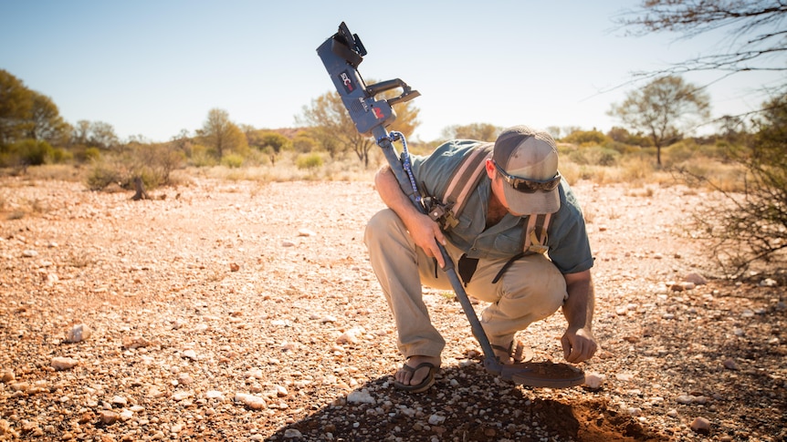 A man crouches on the ground with a metal detector.
