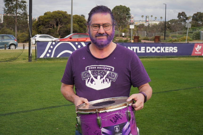 A man in Fremantle Dockers gear holds a purple drum. 
