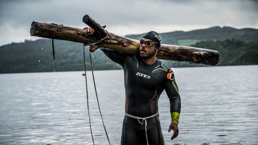 Ross Edgley stands in the water holding a tree trunk.