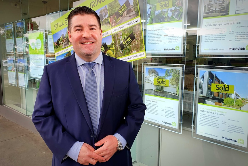 A man in a suit smiles as he stands in front of posters advertising properties for sale.