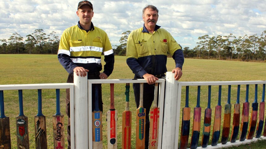 Two men in high vis standing behind a fence made of cricket bats