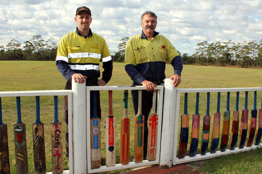 Two men in high vis standing behind a fence made of cricket bats